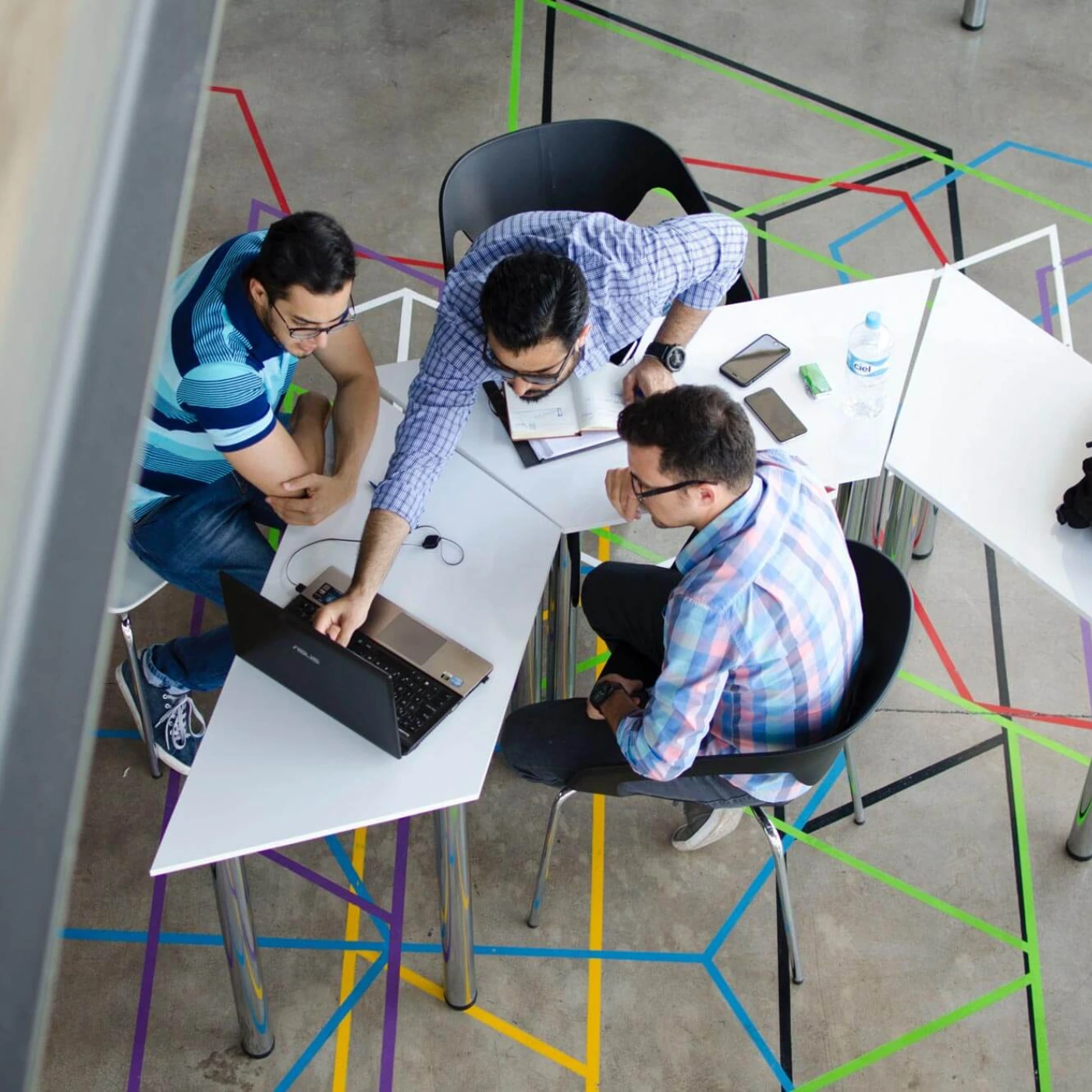 Symbolic image of Abacus Experten GmbH at Abacus alpha: Overhead view of three men working on a laptop in a modern, colorful office with geometric lines on the floor.